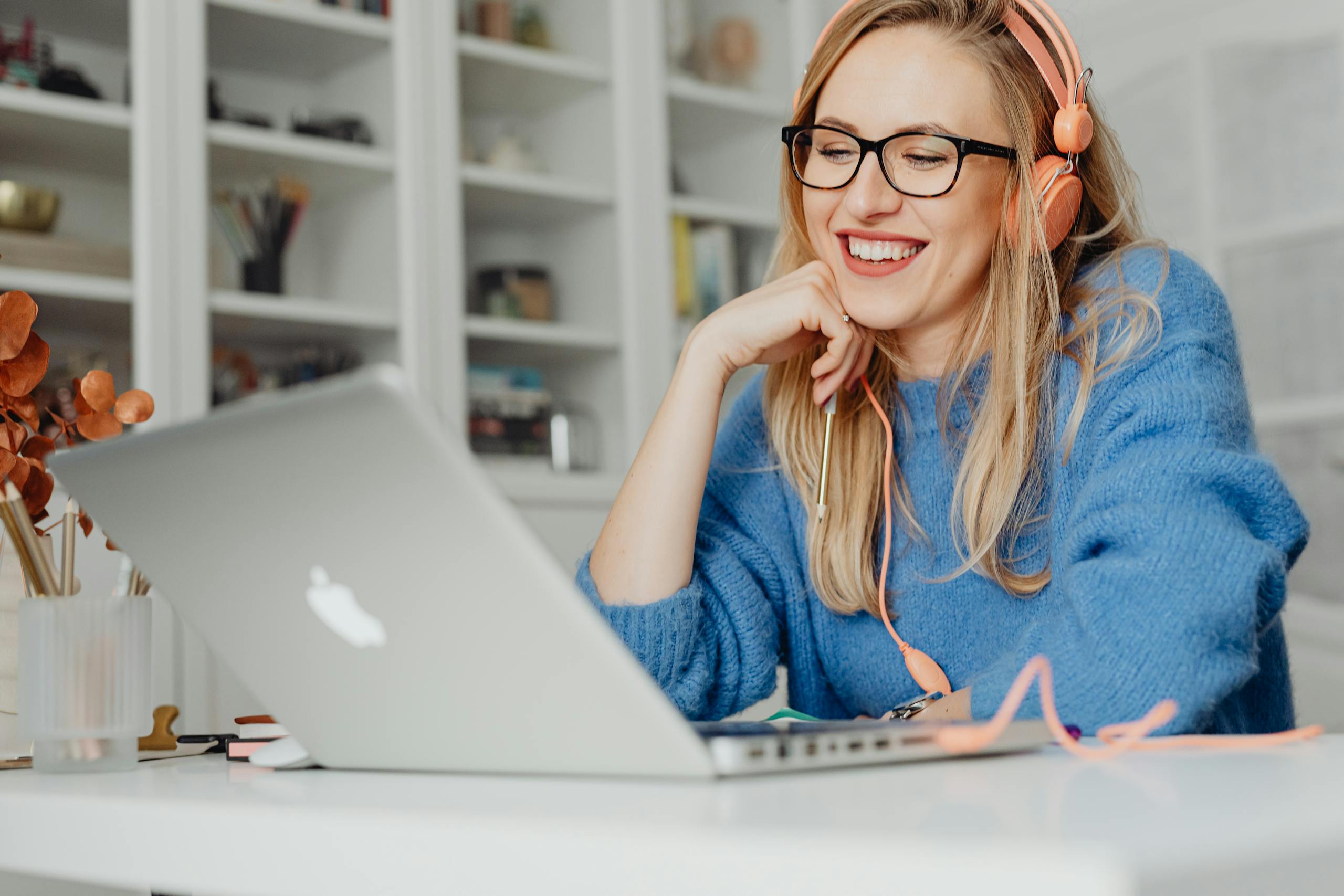 Smiling woman wearing headphones and glasses, working on a laptop in a bright indoor setting.